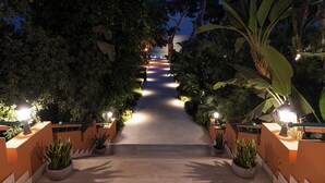 A pathway flanked by potted plants and illuminated by wall-mounted lights, leading towards a body of water under a night sky. - Forte Village Resort - Il Castello (Pula)