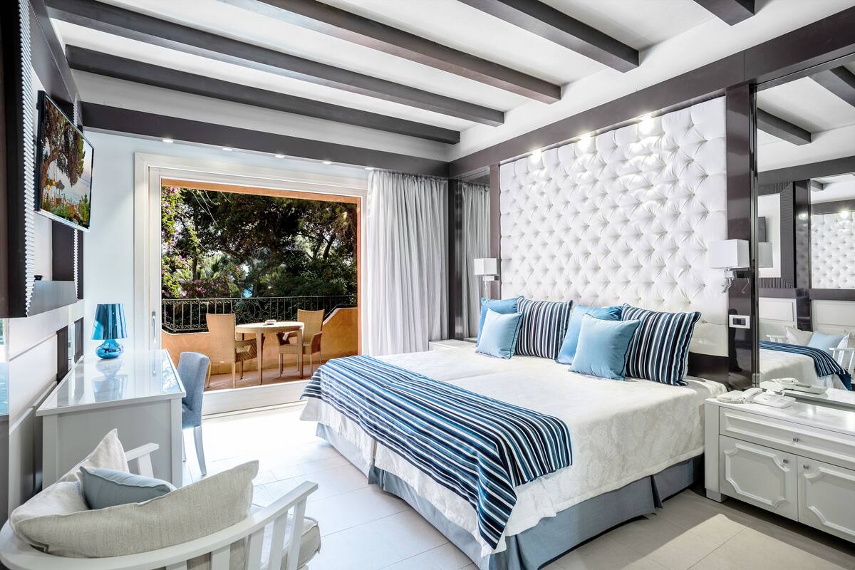 A modern hotel room with a large bed, a balcony with a view of greenery, a small sitting area with a table and chairs, and a bathroom with a large mirror and a vanity.