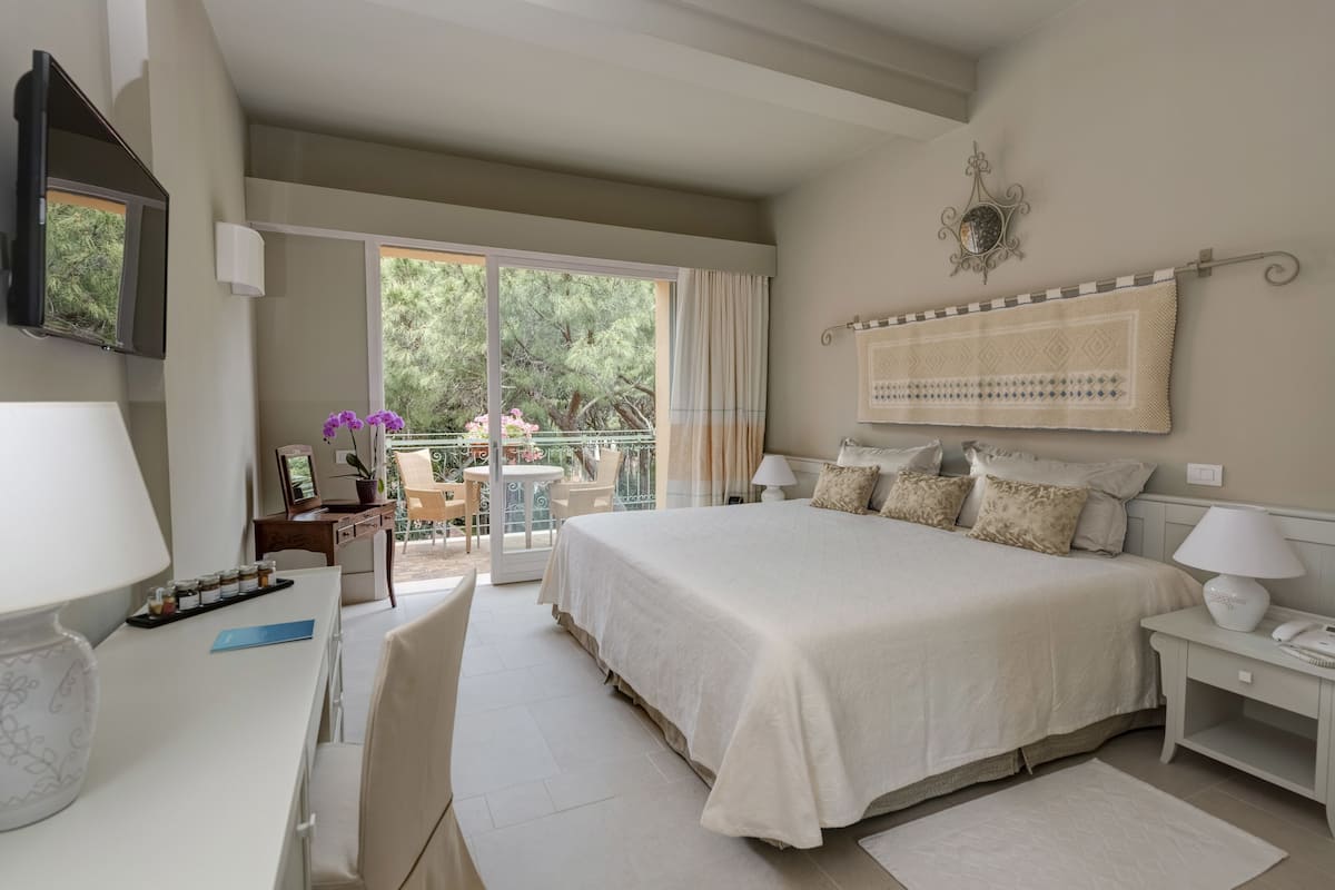 A hotel room with a large bed, a desk with a lamp, a chair, a TV, and a balcony with a table and chairs.