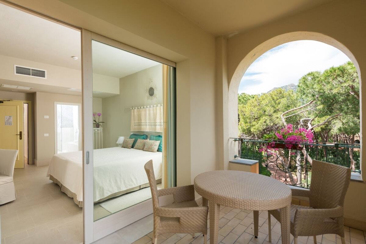 A bedroom with a bed, a balcony with wicker furniture, and a view of trees.