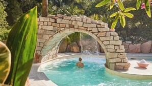 A stone bridge over a pool with a person swimming, surrounded by greenery. - Forte Village Resort - Il Castello (Pula)