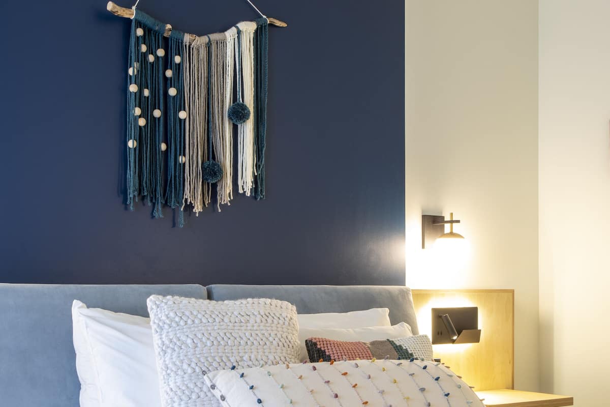 A modern bedroom with a bed, pillows, a wall-mounted macrame piece, and a bedside table with a lamp.
