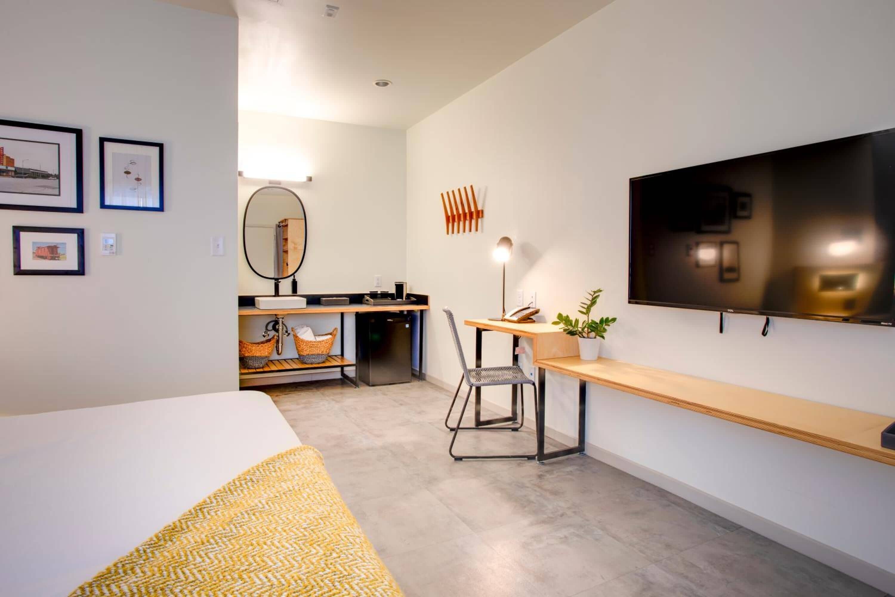 Een moderne hotelkamer met een bed, een bureau met stoel, een flatscreen televisie, een spiegel en een kleine kitchenette.