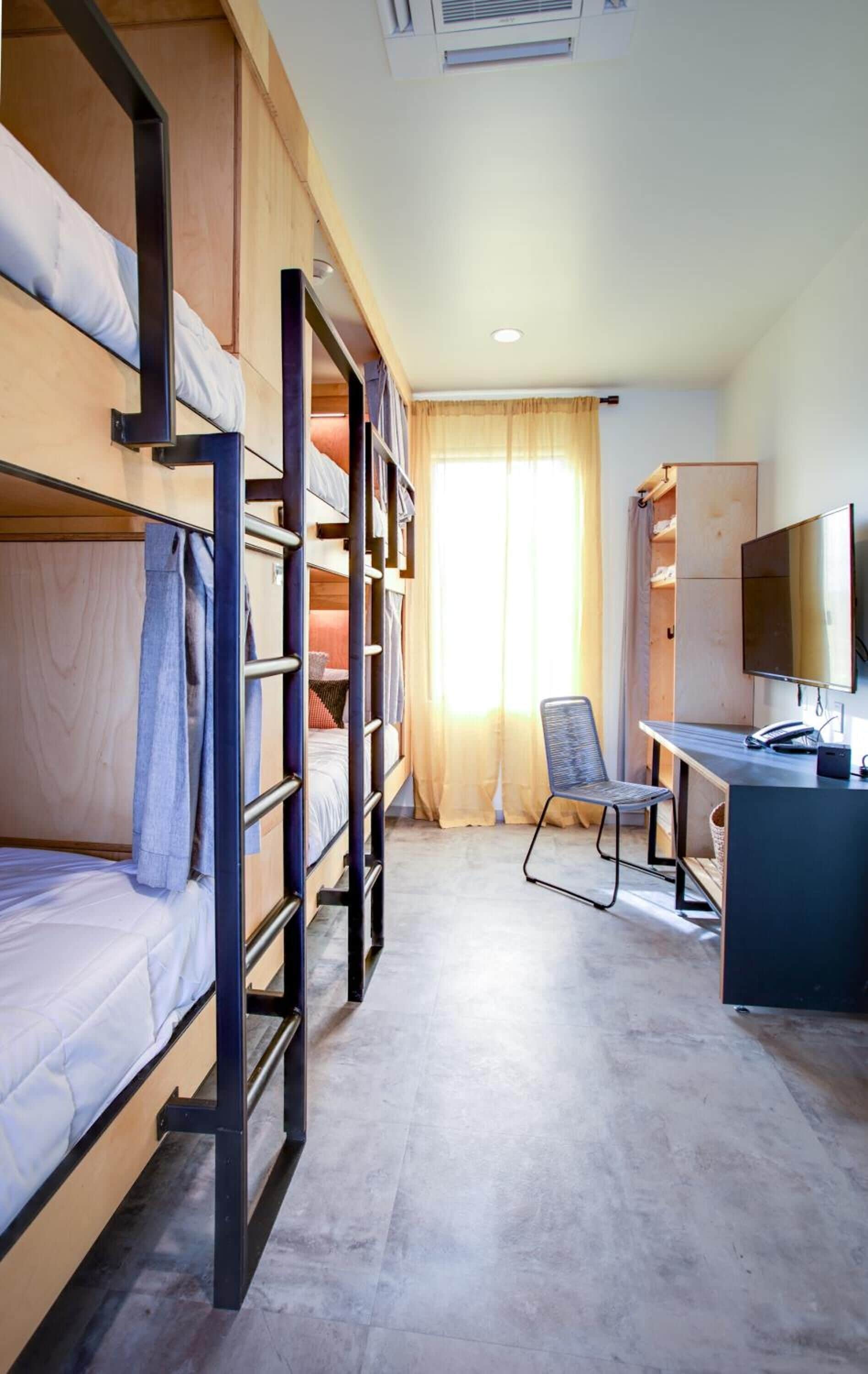 Quarto estilo dormitório com beliches, escrivaninha, cadeira, televisão e guarda-roupa.