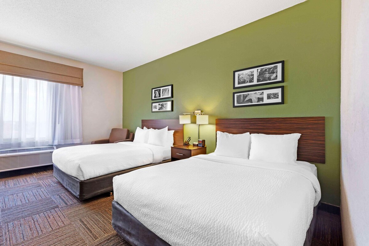 A hotel room with two beds, a large window with curtains, a wall-mounted lamp, and framed pictures.