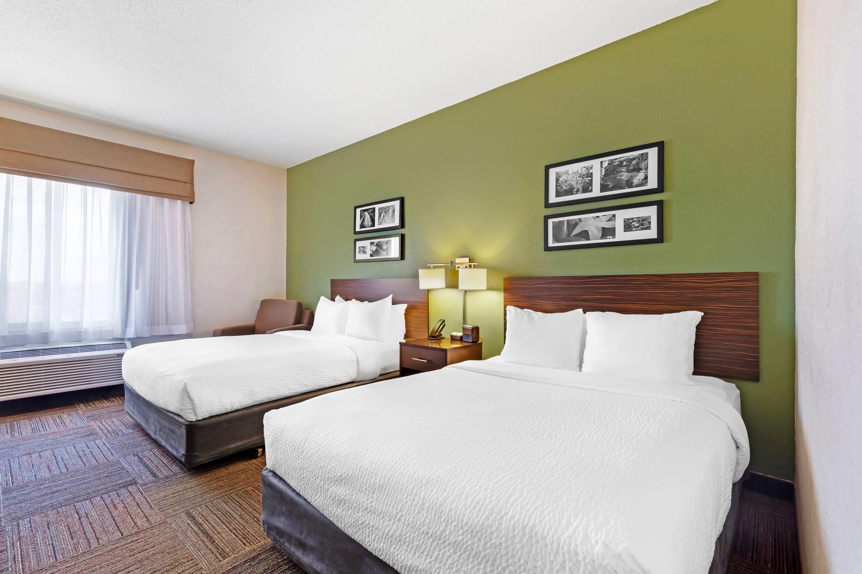 A hotel room with two beds, a bedside table with a lamp, a chair, and a window with curtains.