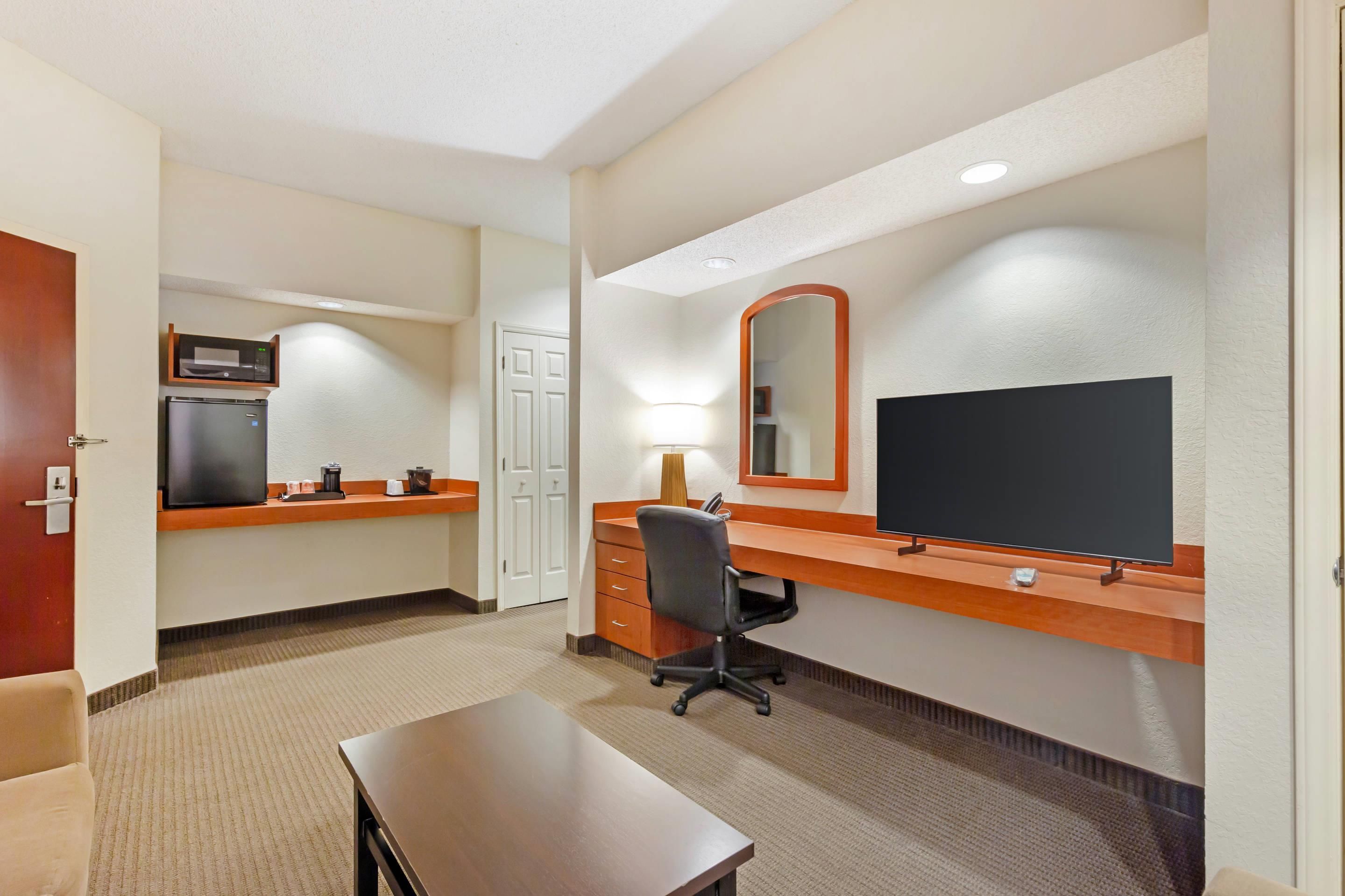 Quarto de hotel com cozinha compacta, escrivaninha com computador, cadeira, cama e televisão.