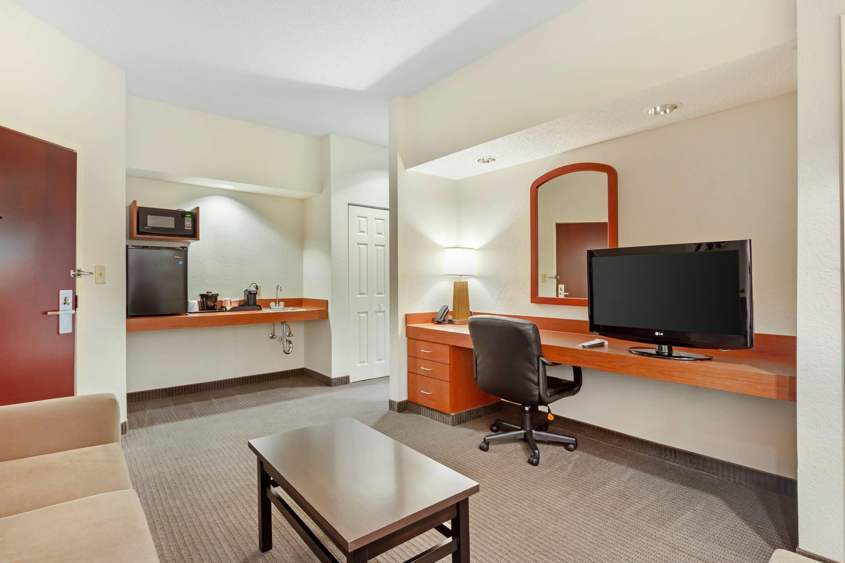 A hotel room with a kitchenette, a desk with a computer and TV, a chair, and a small table.