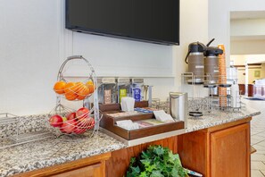 Een ontbijtbuffet in een hotel met fruitmanden, koffiezetapparaten en een flatscreen televisie.