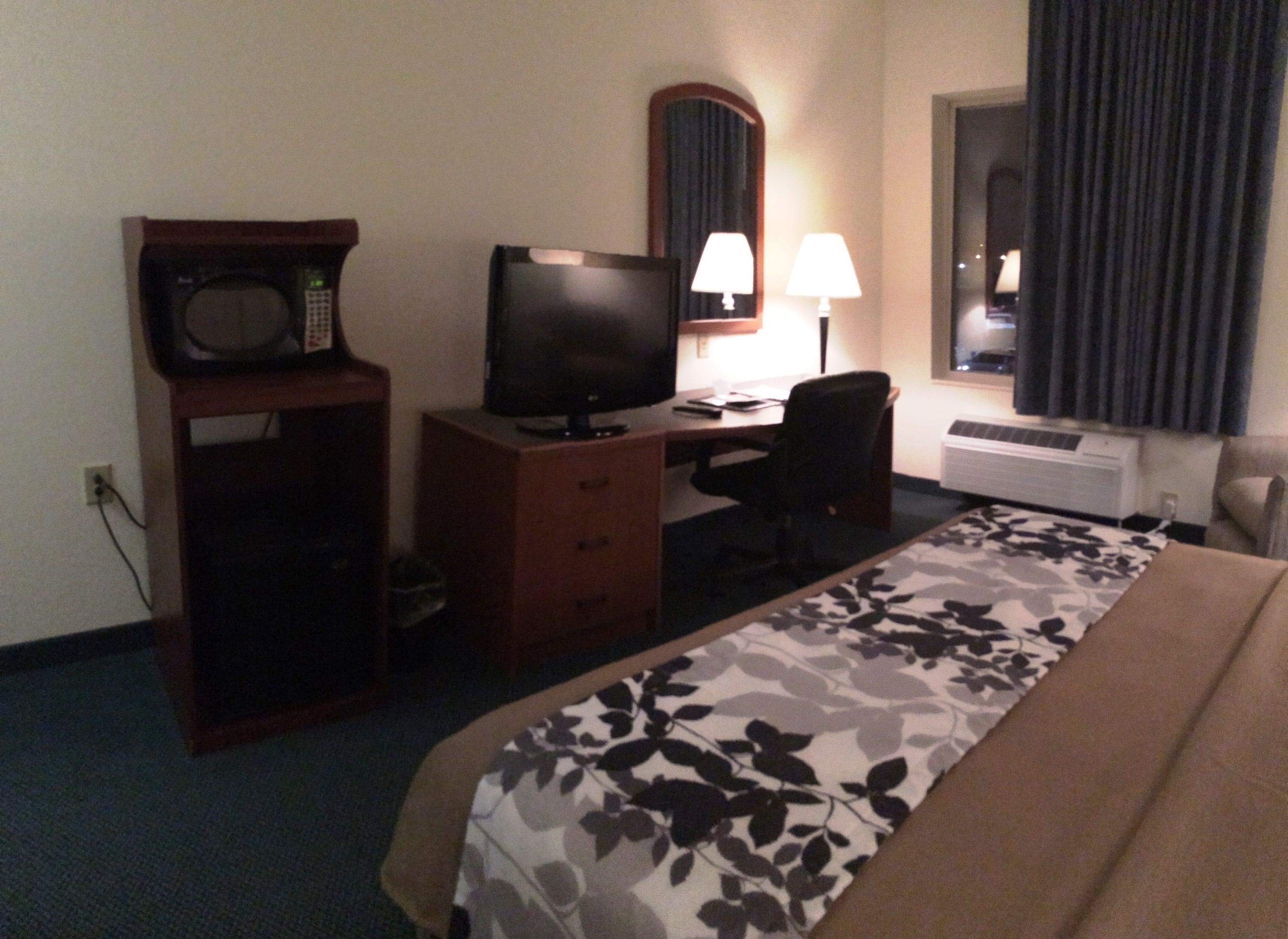A hotel room with a bed, a desk with a computer, a television, a microwave, a chair, and a window with curtains.