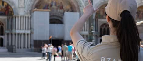 A person wearing a cap and a shirt with 'BZA HOTELS' text, pointing towards a historic building with ornate architecture and a crowd in the foreground.