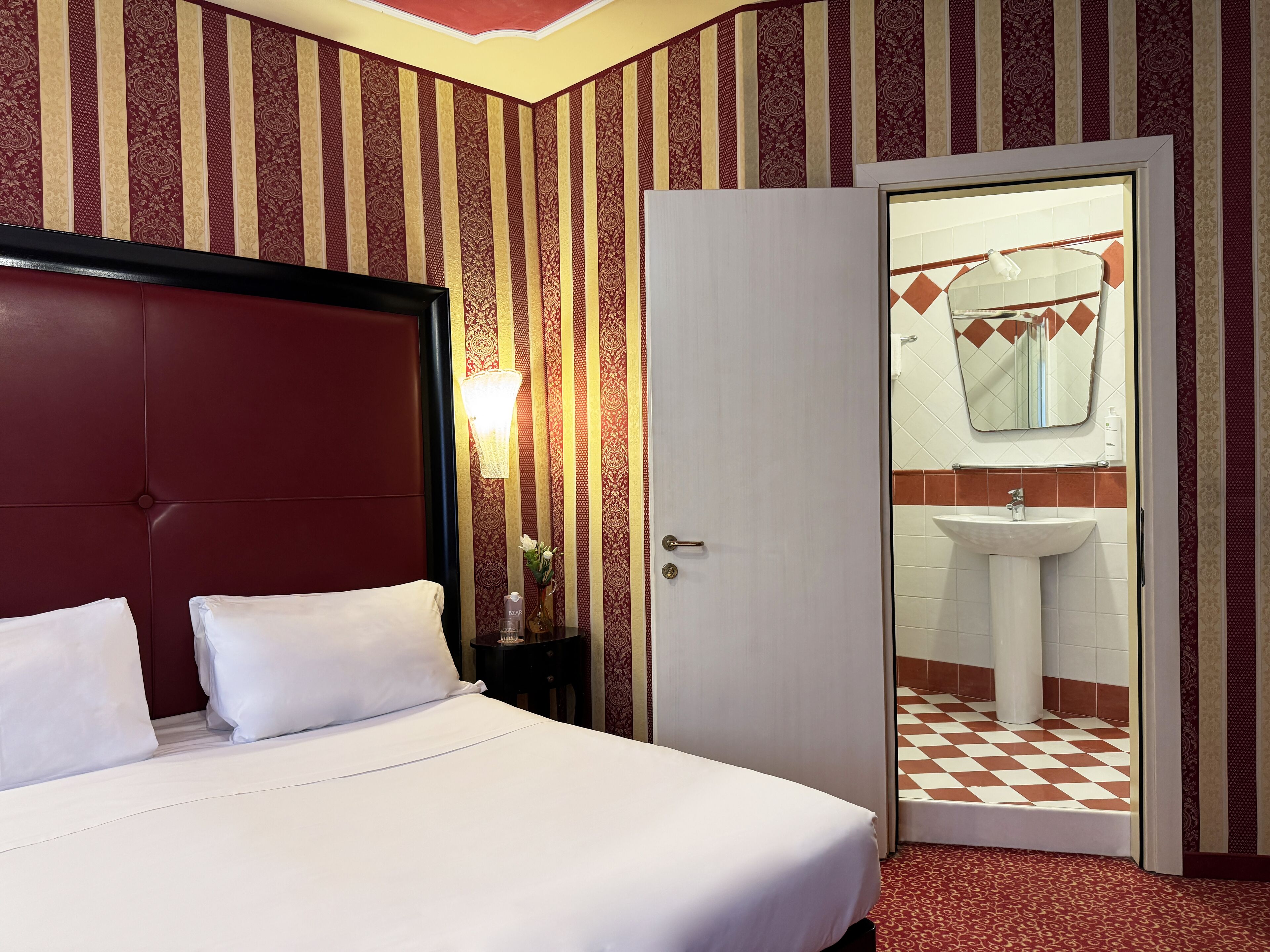 Habitación de hotel con una cama grande, una mesita de noche, una lámpara y una puerta que da a un baño con lavabo y espejo.