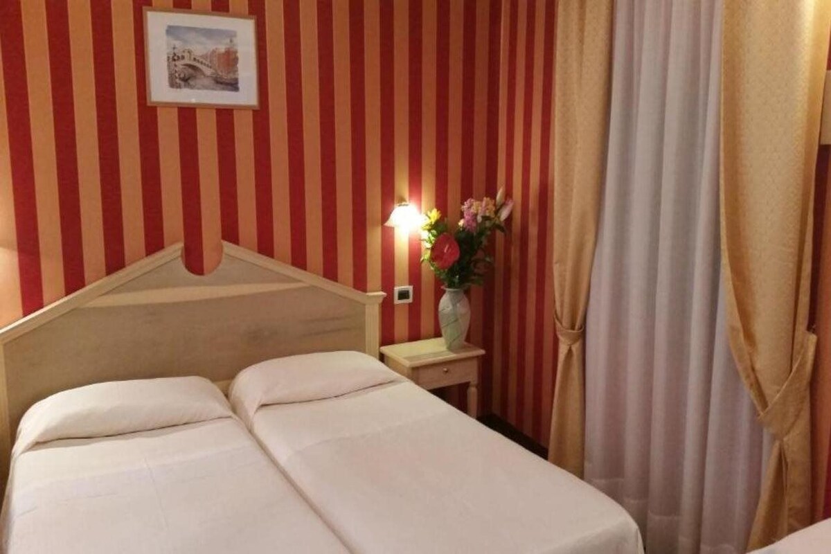 A hotel room with a striped red and white wall, a bed with white linens, a nightstand with a vase of flowers, and a lamp.
