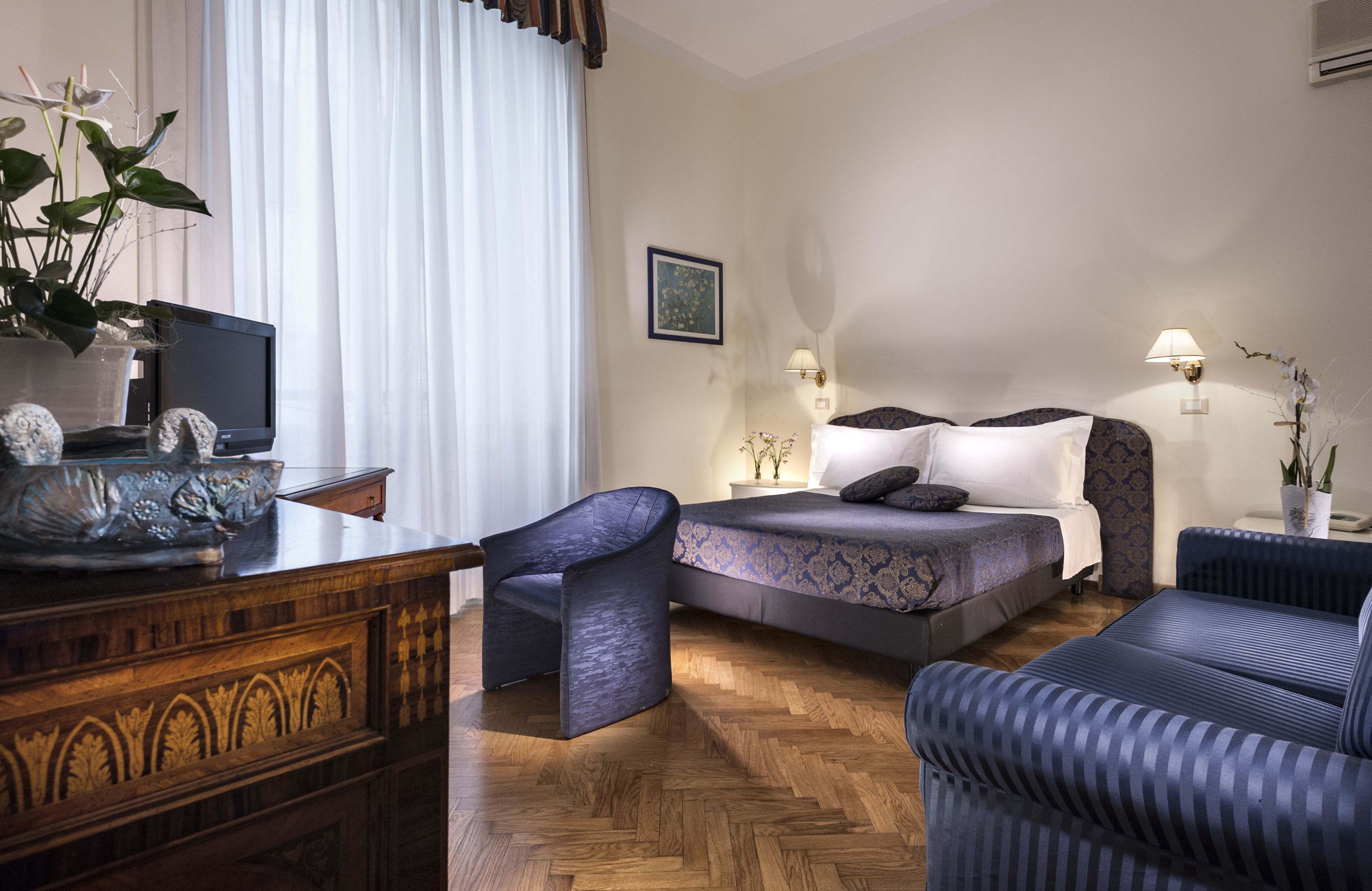 Habitación de hotel con escritorio de madera, televisor, cama con ropa de cama estampada, dos sillas y una planta en maceta.