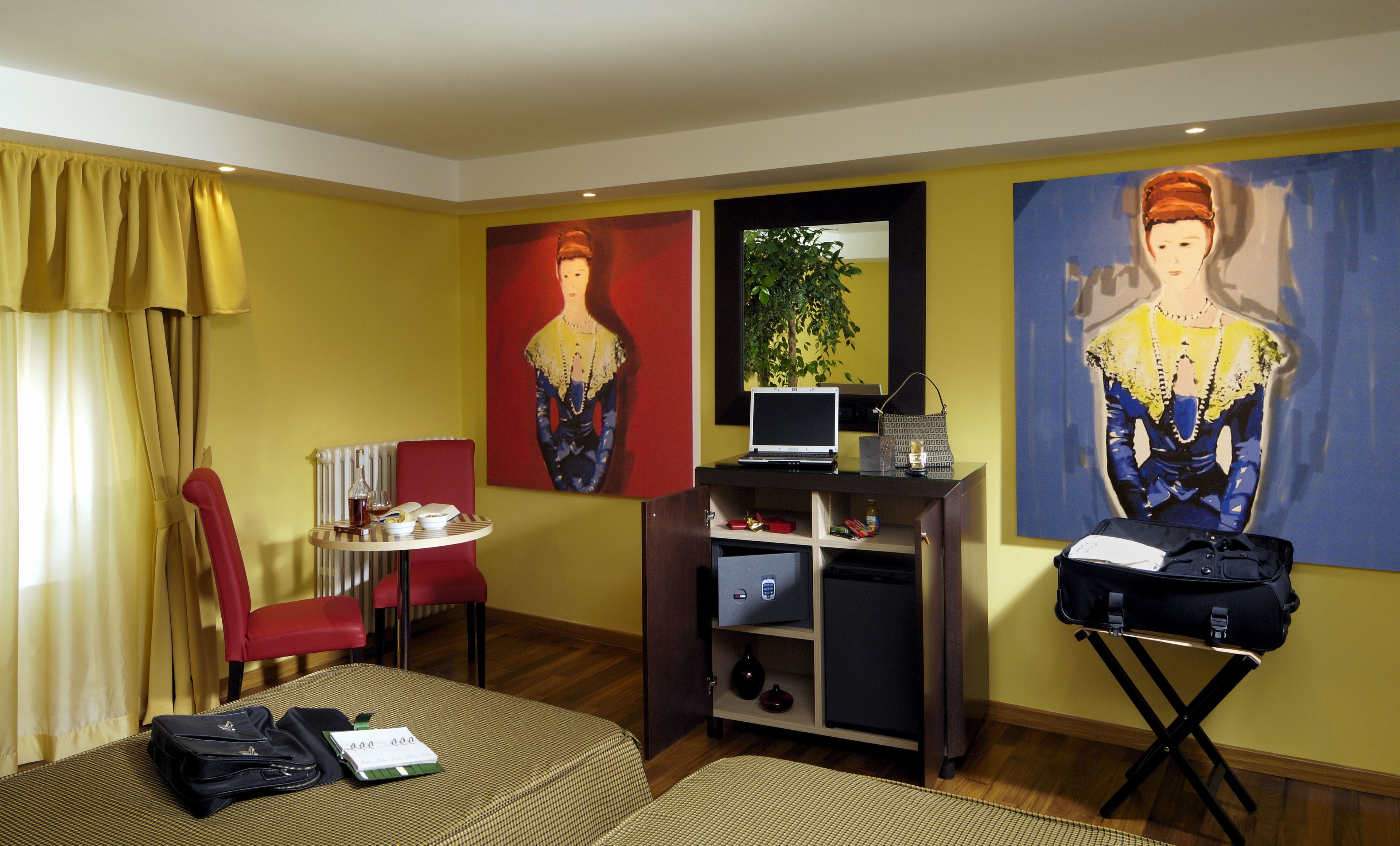 Une chambre d’hôtel avec un lit, un bureau avec un ordinateur portable, une chaise, une petite table avec une bouteille et des verres, et deux tableaux accrochés au mur.