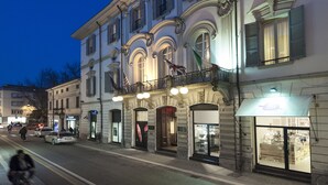 Un edificio histórico con arquitectura ornamentada, que incluye un balcón con barandillas y banderas, y locales comerciales en la planta baja.