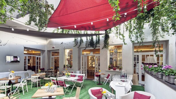 Un patio al aire libre con mesas y sillas, un toldo rojo y follaje.