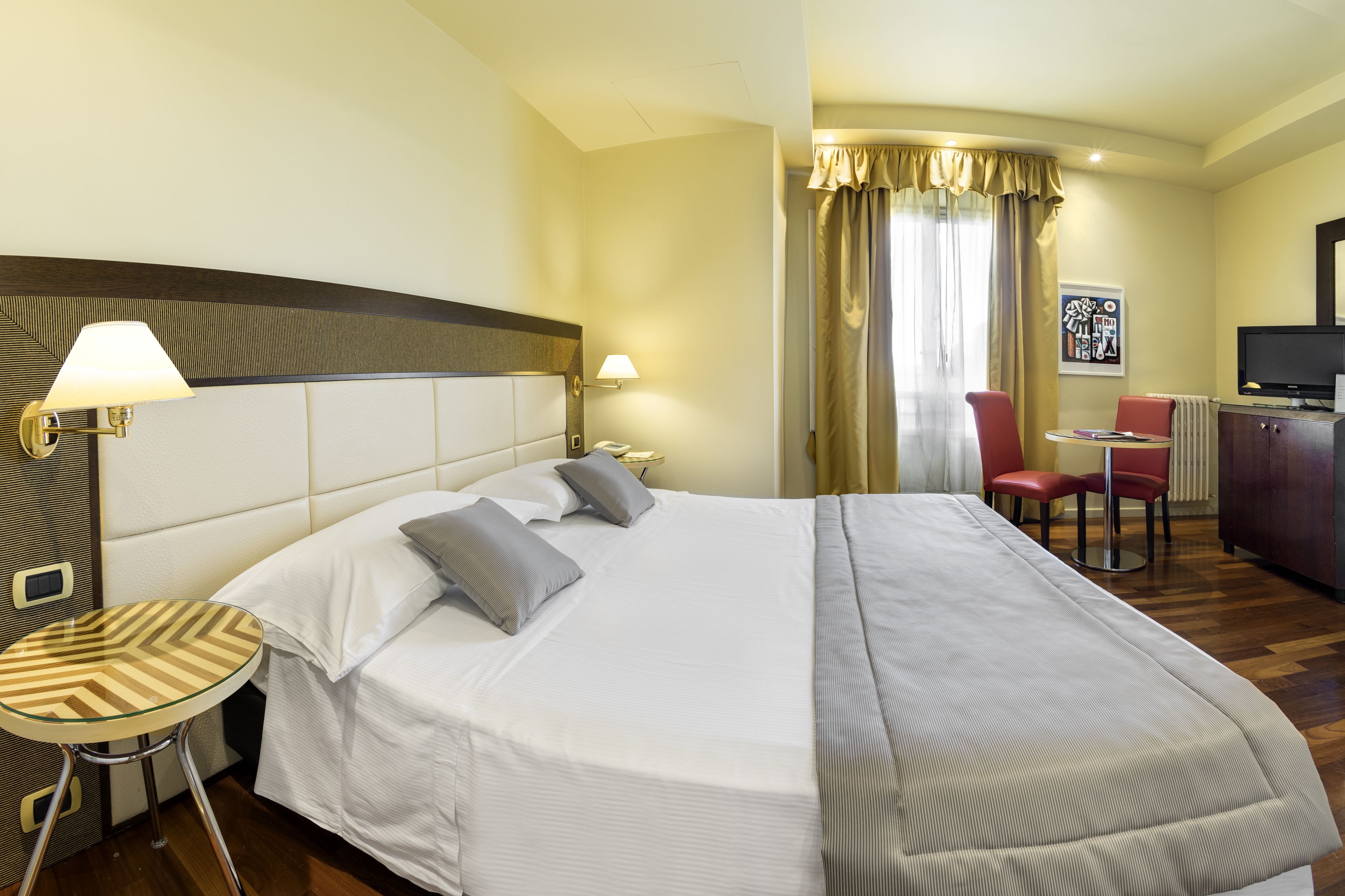Una habitación de hotel con una cama grande, una mesita redonda pequeña, dos sillas, un escritorio con televisor y una ventana con cortinas.