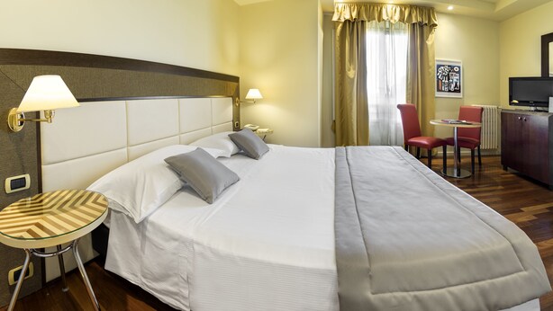 A hotel room with a large bed, a small round table, two chairs, a desk with a television, and a window with curtains. - Vittoria (Faenza)