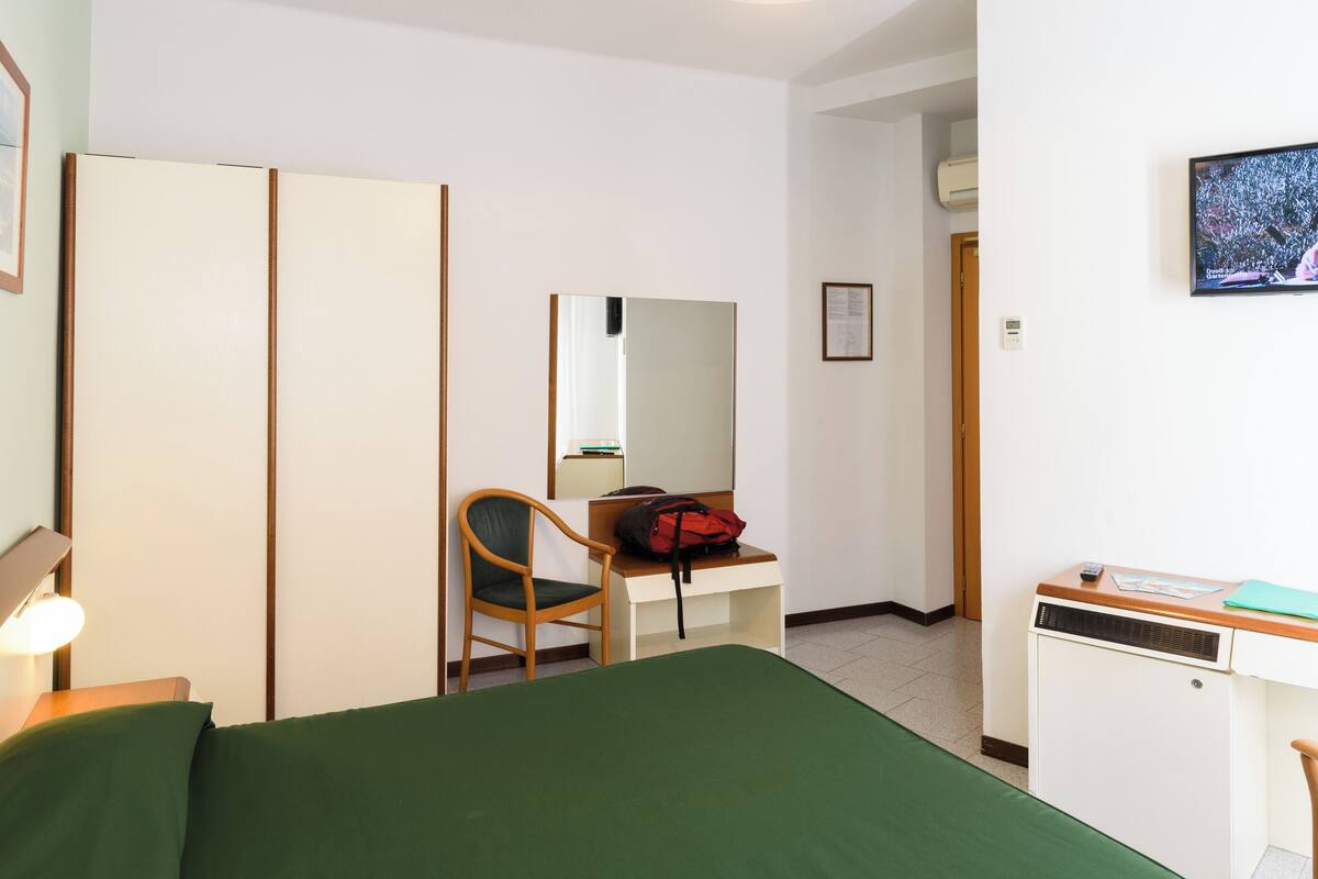 Une chambre d’hôtel comprenant un lit, un bureau avec une chaise, un miroir, une armoire et un petit radiateur.