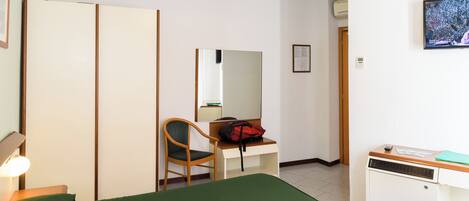 A hotel room with a bed, a desk with a chair, a mirror, a wardrobe, and a small heater.