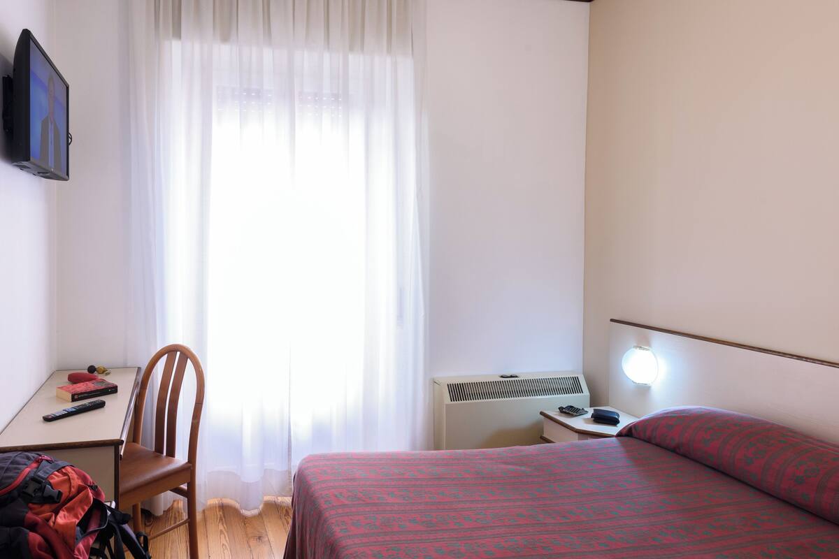 Une chambre d’hôtel comprenant un lit, un bureau avec une chaise, un téléviseur fixé au mur, une fenêtre avec des rideaux et une lampe de chevet.