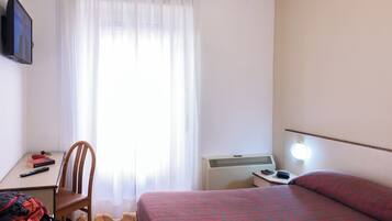 A hotel room with a bed, a desk with a chair, a TV mounted on the wall, a window with curtains, and a bedside lamp.