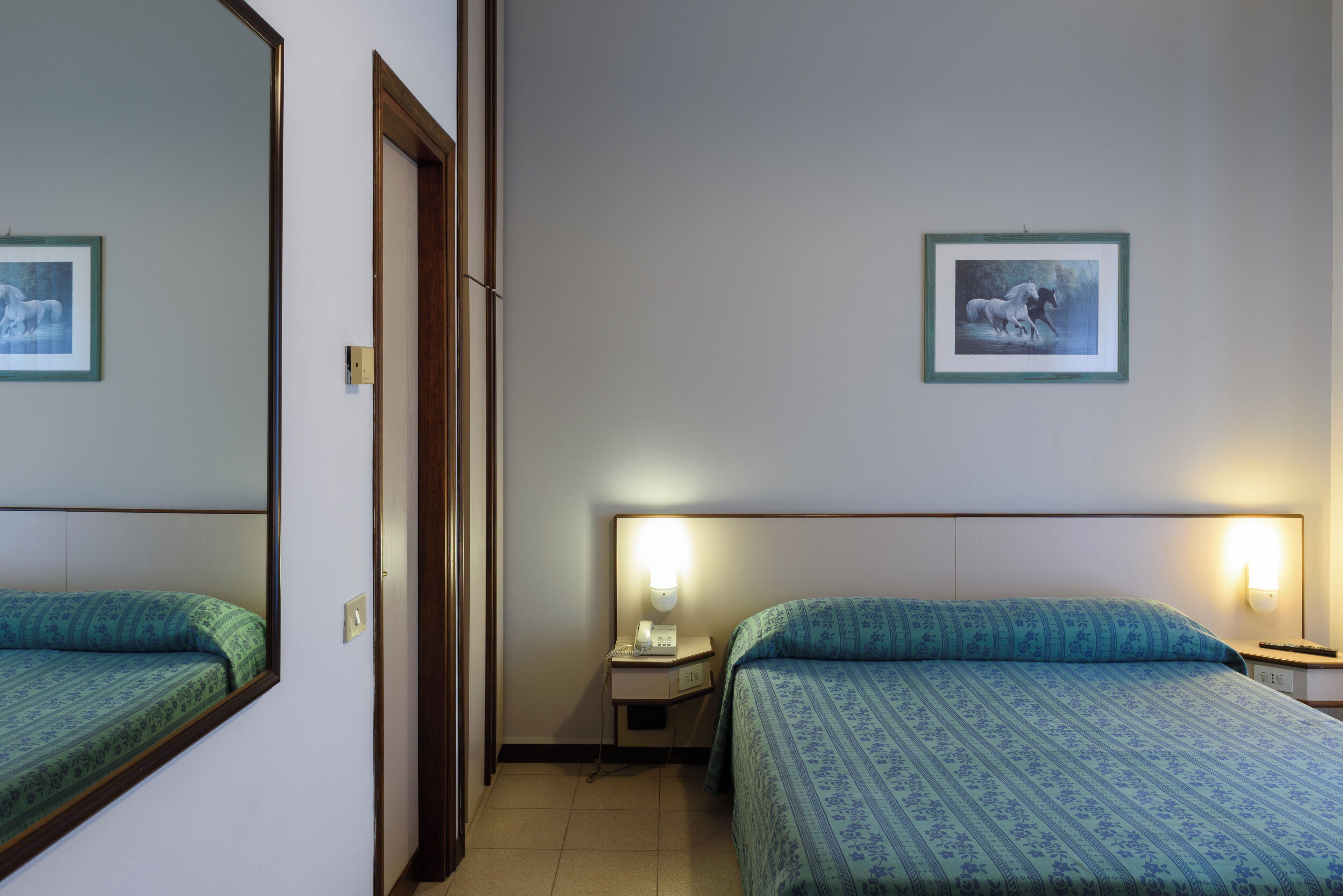 A hotel room with two beds, a nightstand with a lamp, and two framed pictures on the wall.