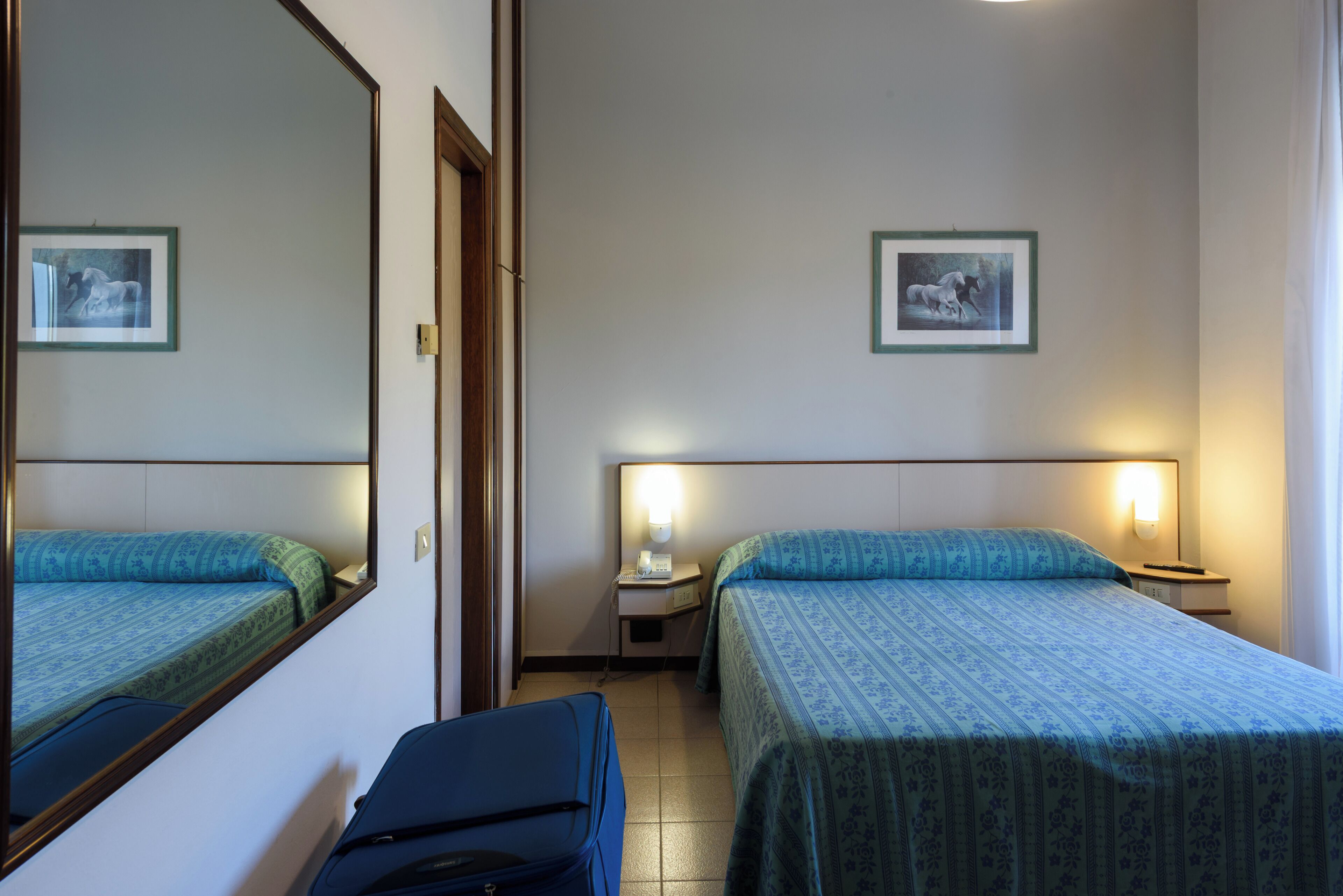 Hotellihuone, jossa on kaksi sänkyä, työpöytä, tuoli, matkalaukku ja peili.