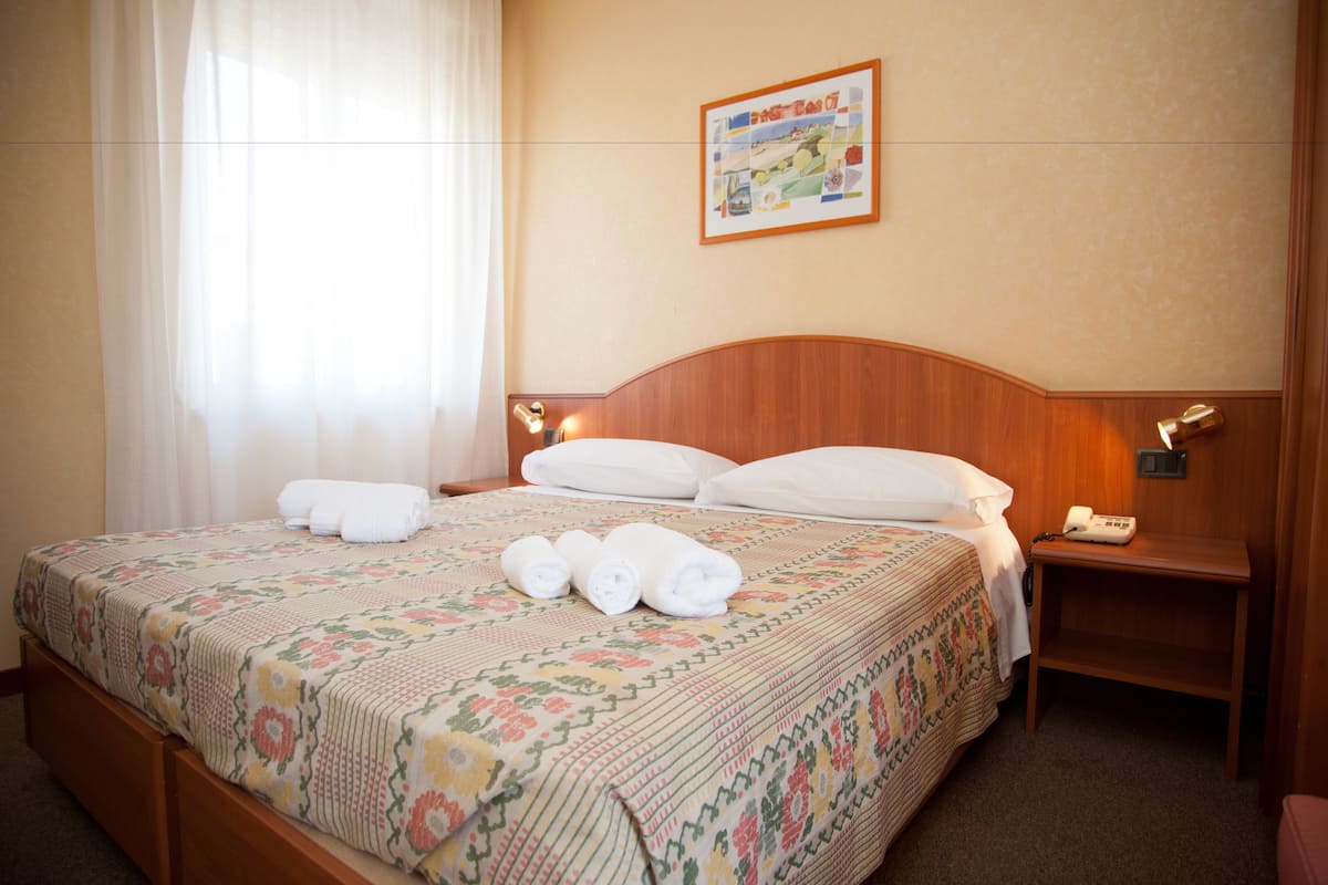 A hotel room with a wooden headboard, a floral-patterned bedspread, neatly folded towels, a bedside table with a lamp, and a framed picture on the wall.
