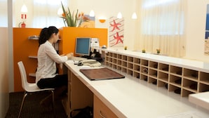 A reception desk with a computer, a telephone, and a plant. There is a wall with a fire extinguisher and a window with curtains. - Hotel Rodia (Maglie)