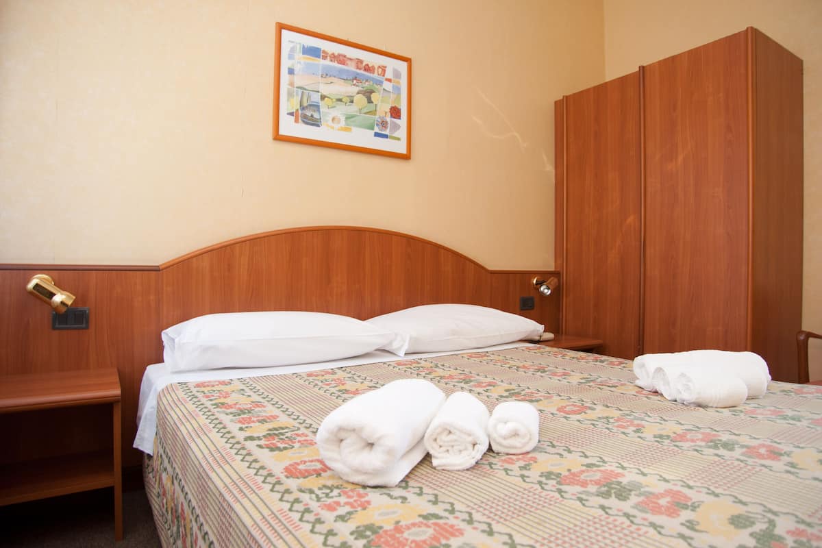 A hotel room with a wooden headboard, a bed with white linens, a wooden wardrobe, and a framed picture on the wall.