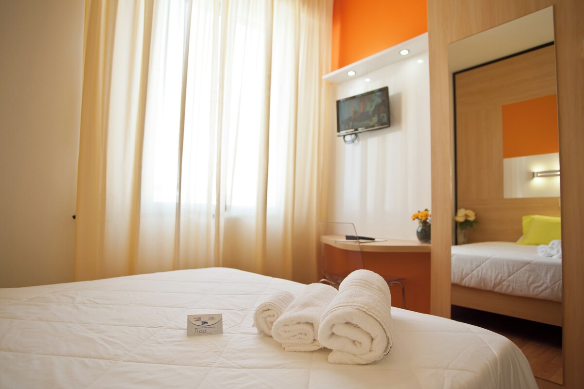 Habitación de hotel con cama, escritorio con computadora, televisor montado en la pared y ventana con cortinas.