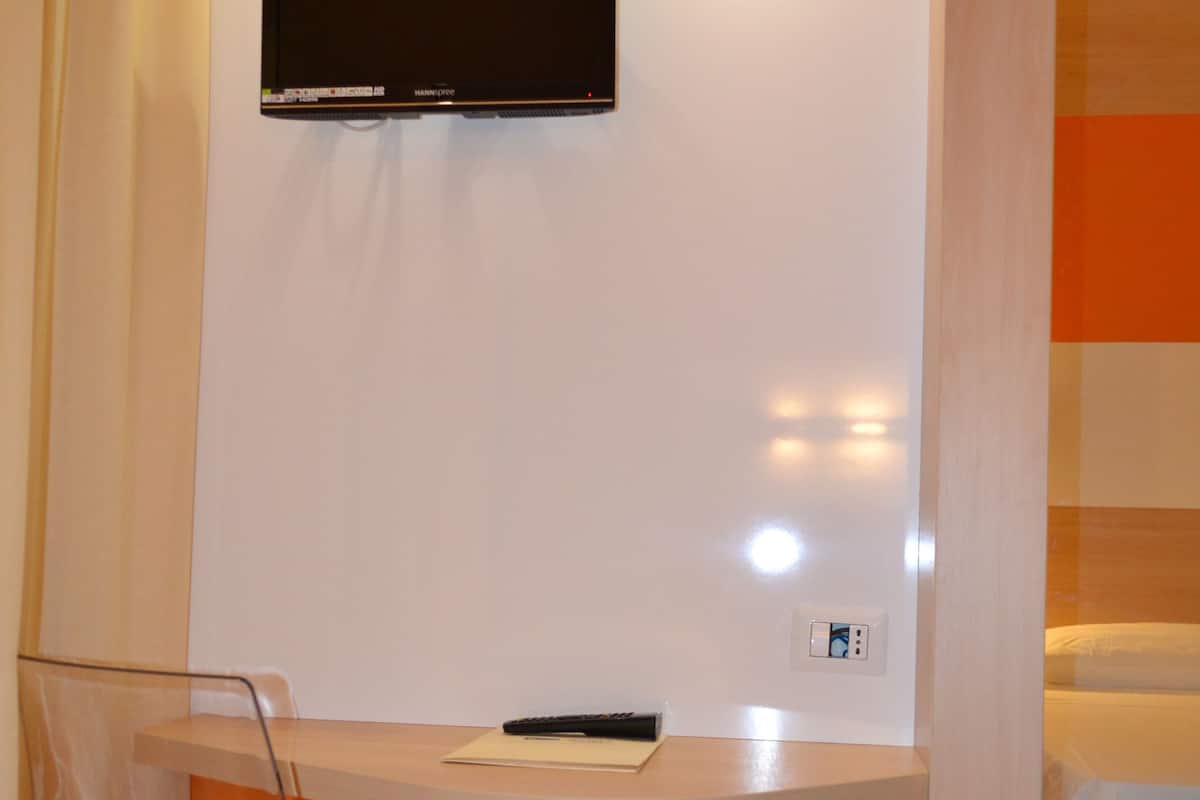 A modern hotel room with a flat-screen TV mounted on the wall, a wooden desk, a clear plastic chair, and a bed with white bedding.