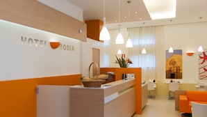 A modern hotel reception area with a light wood reception desk, a seating area with orange and white chairs, and a large window with curtains. - Hotel Rodia (Maglie)