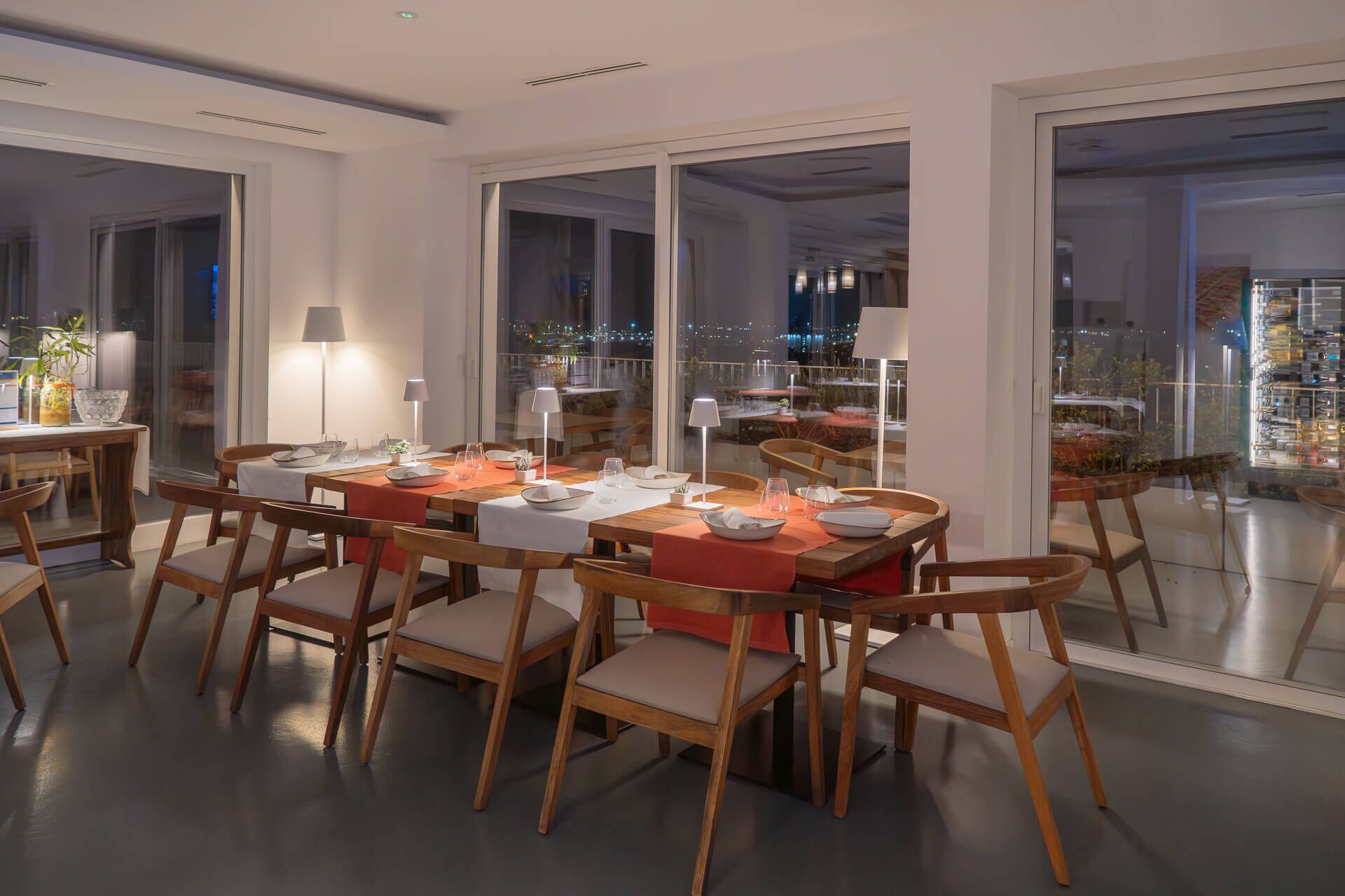 A dining area with a large table set for six, surrounded by wooden chairs with cushioned seats. The room has large glass doors leading to an outdoor terrace with a city view.