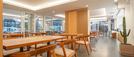A modern dining area with wooden tables and chairs, large windows, and a cactus plant.