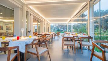 A modern restaurant with wooden tables and chairs, large windows offering a view of the outdoors, and a ceiling with linear lighting.