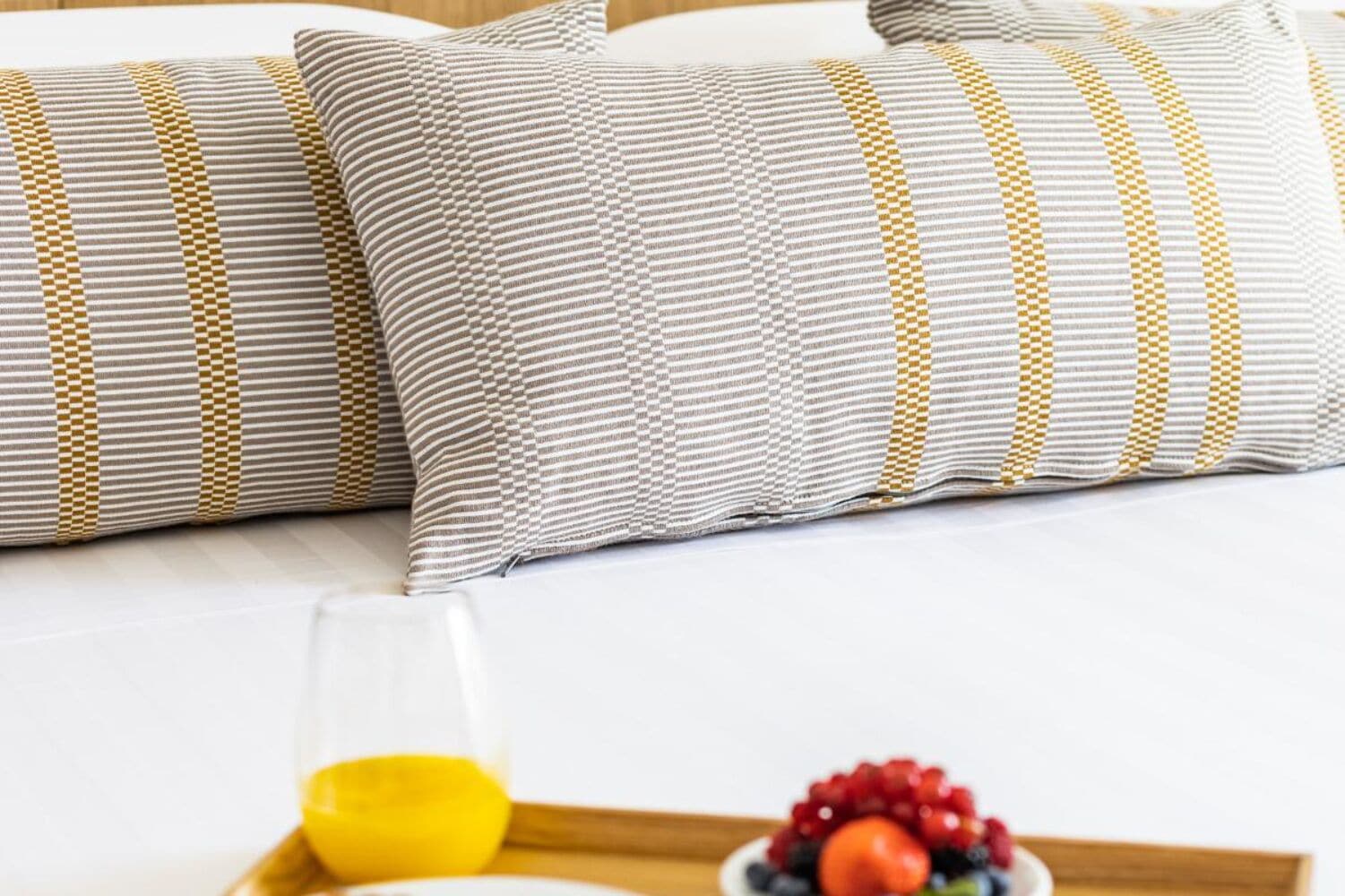 Ein ordentlich bezogenes Bett mit gestreiften Kissen und einem Tablett mit Orangensaft und einer Schale Obst.
