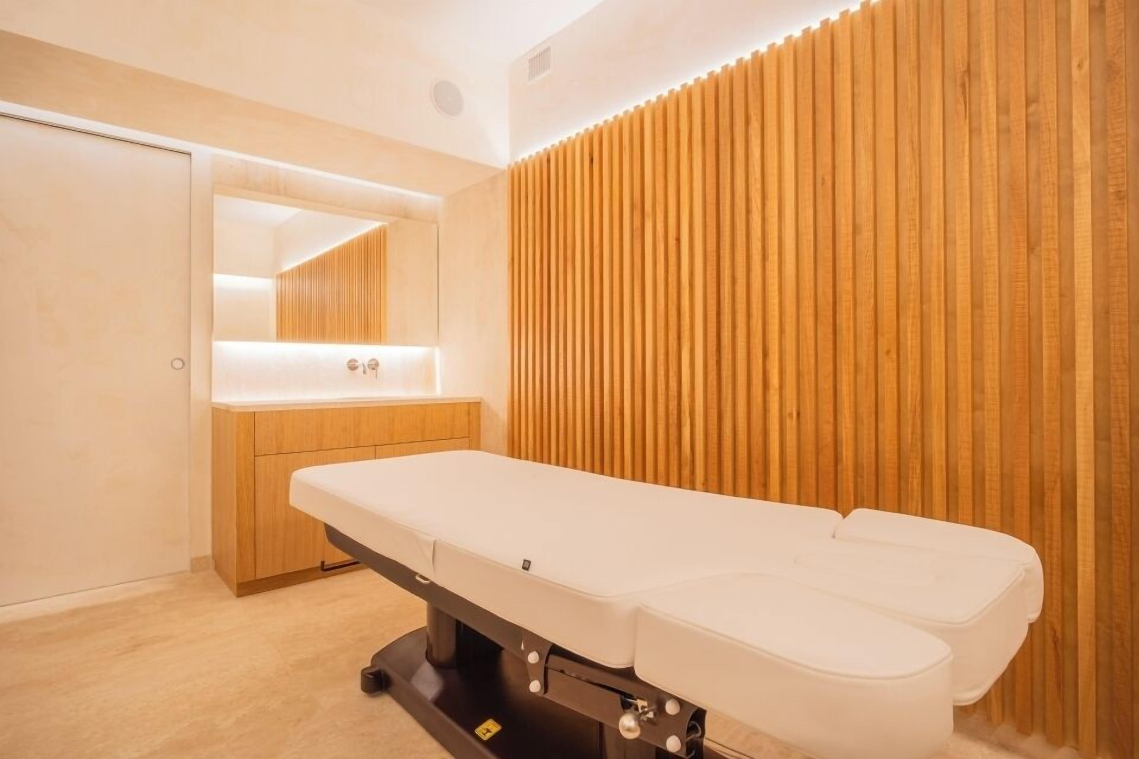 Un moderno consultorio médico con pared de paneles de madera, una camilla blanca y un mueble de madera con espejo.