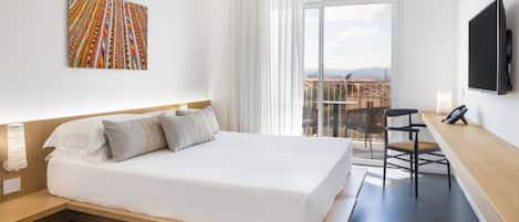 A modern hotel room with a large bed, a desk with a chair, a TV, and a balcony with a city view.