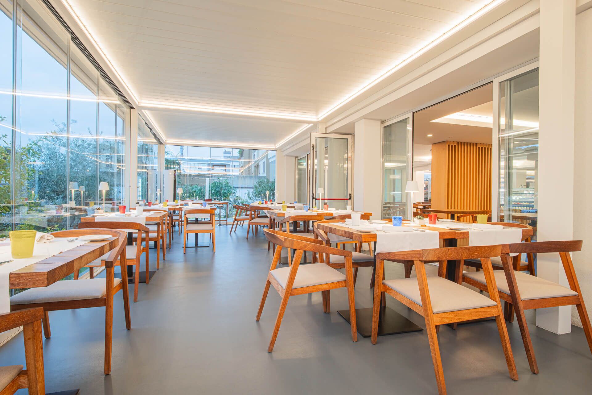 A spacious indoor dining area with wooden tables and chairs, large windows offering views of greenery, and a modern ceiling design.