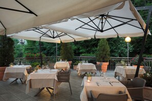 Zona pranzo all'aperto con tavoli e sedie sotto grandi ombrelloni, circondata da piante in vaso e un parapetto.