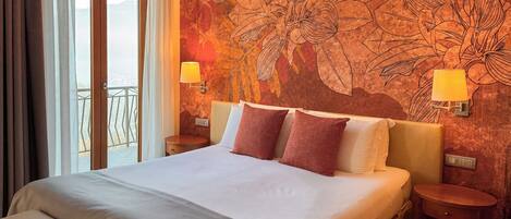 A hotel room with a large bed, two bedside lamps, a nightstand, a wooden floor, and a view of the outdoors.