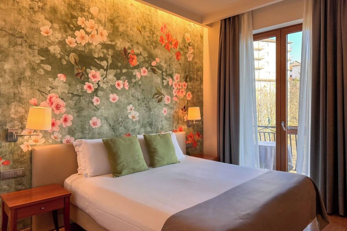 Een hotelkamer met een groot bed, twee nachtkastjes, een wand met bloemenbehang en een raam met gordijnen.