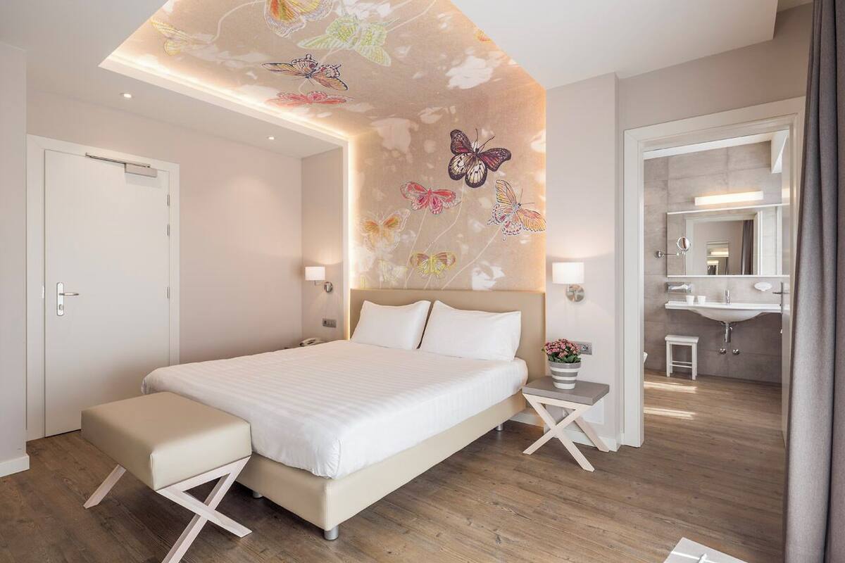 Een moderne slaapkamer met een bed, nachtkastjes, een bankje, een muur met vlinderkunst en een badkamer die door een open deur zichtbaar is.