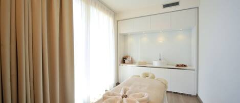 A spa room with a massage table, towels, a window with curtains, and a small kitchenette area.