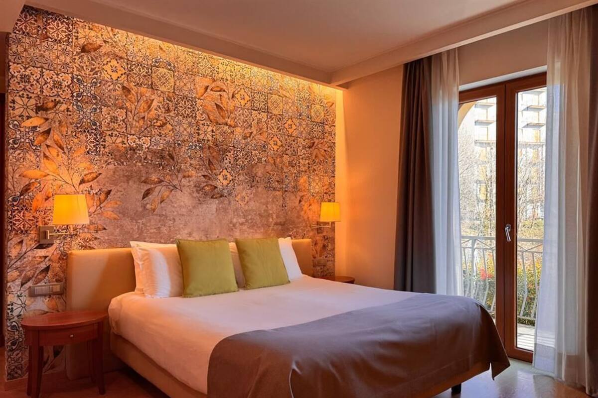 Hotelkamer met een groot bed, twee bedlampen, een wand met stenen motief en een balkon met gordijnen.