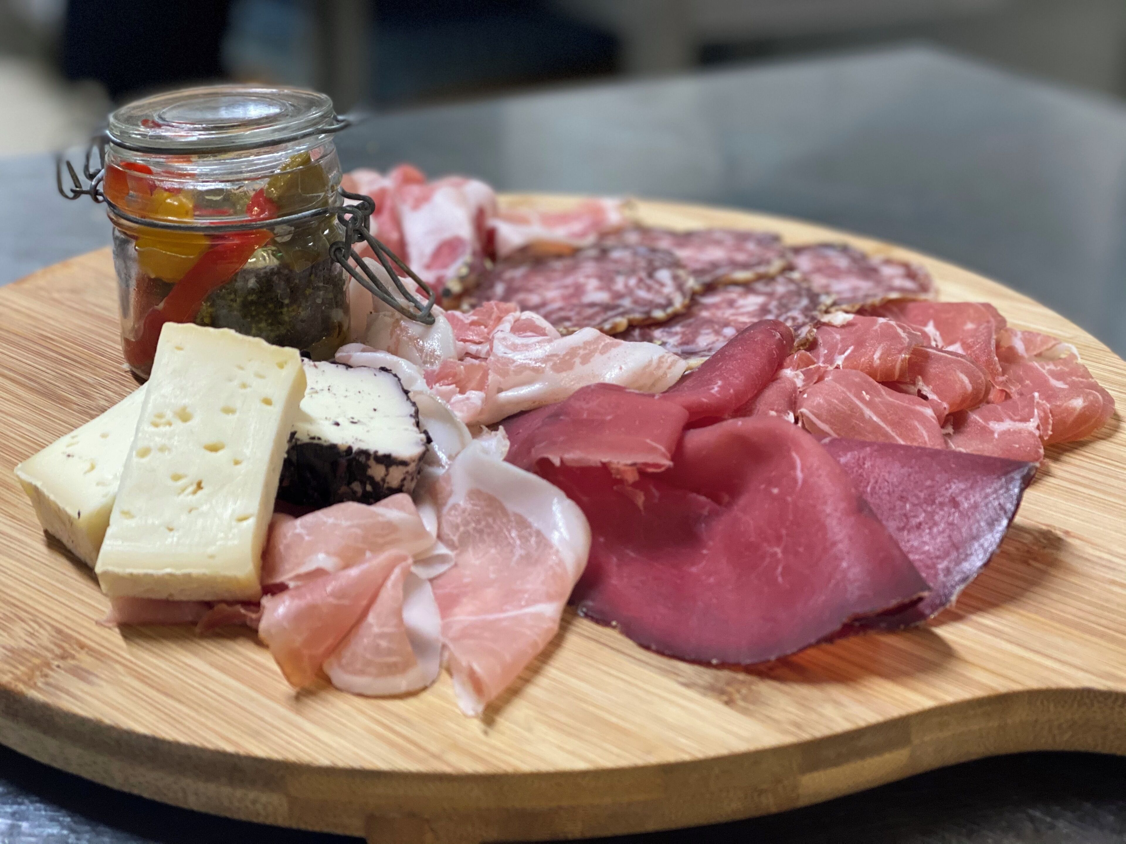 A wooden board with various meats, cheese, and a jar of pickles.