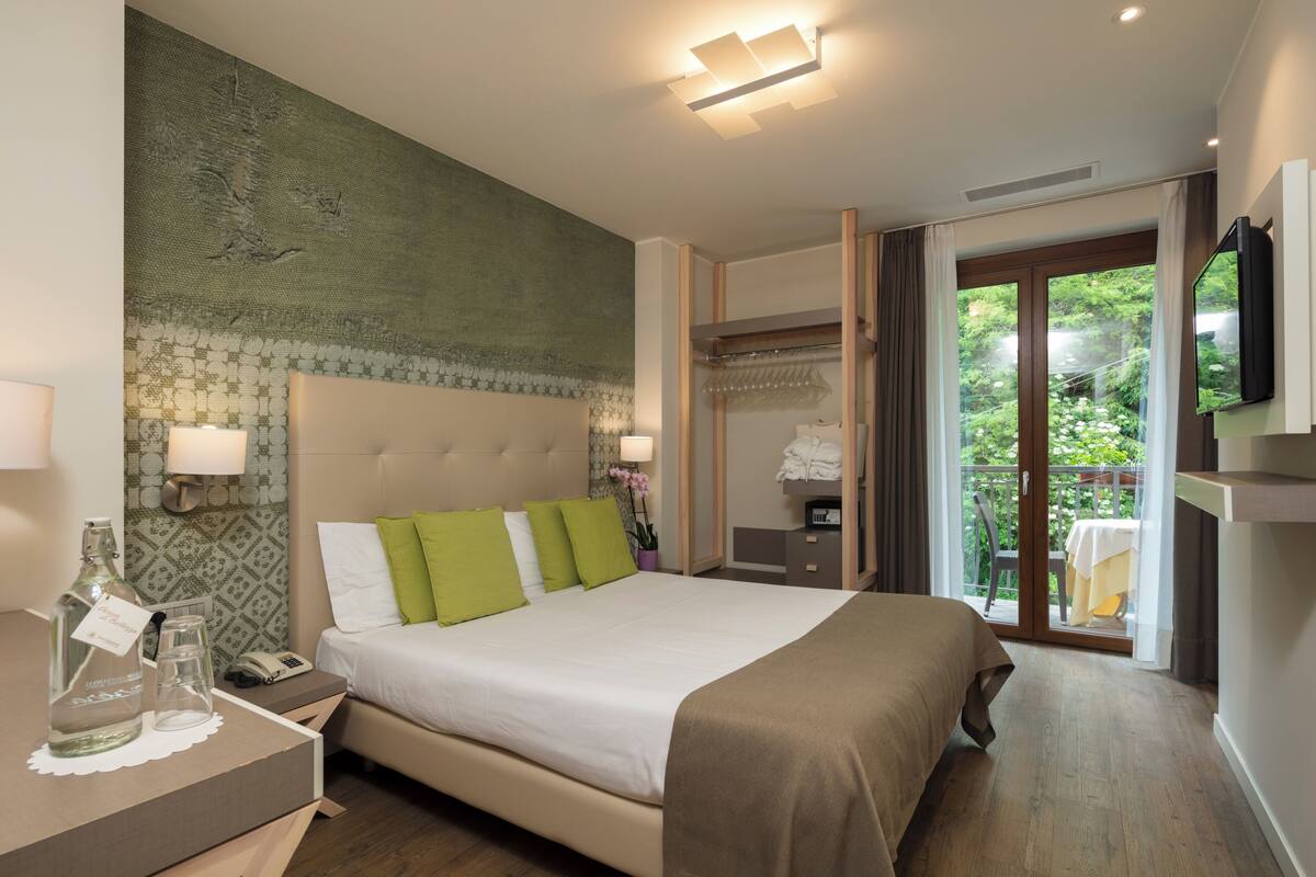 Een hotelkamer met een bed, nachtkastje, lamp en uitzicht op groen buiten het raam.